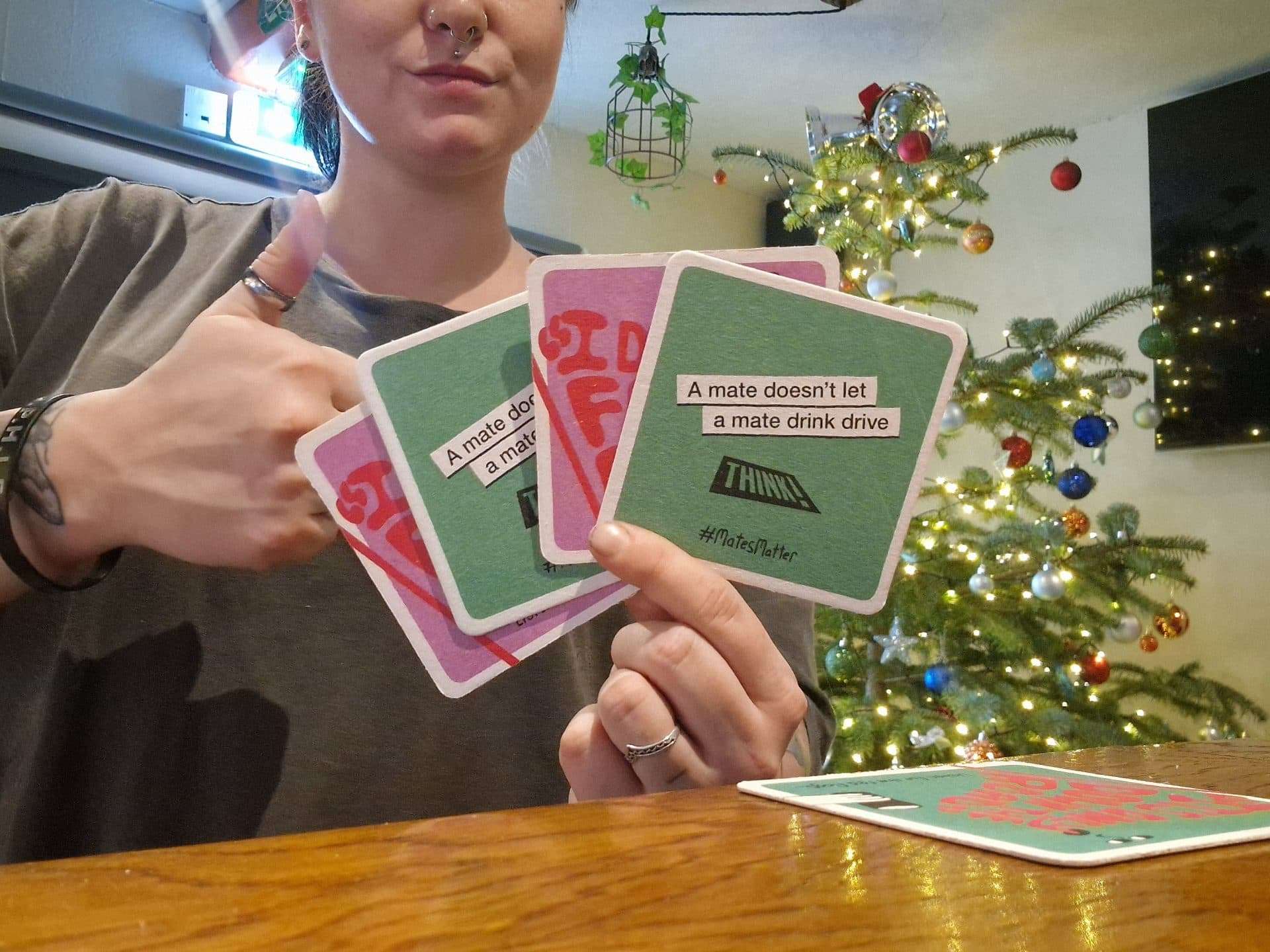 Person giving a thumbs up while holding THINK! beer mats reading 'A mate doesn't let a mate drink drive' in a pub with a Christmas tree