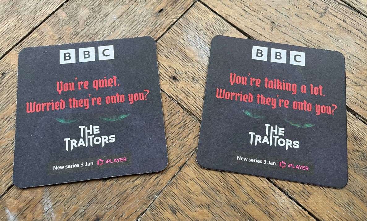 Two black BBC The Traitors beer mats reading 'Worried they're onto you?' promoting the new series on iPlayer, on a wooden table