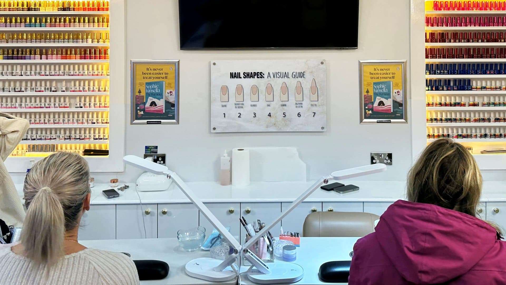 Two framed posters for Sophie Kinsella's novel The Burnout on the wall of a nail salon, with customers seated at the nail bar