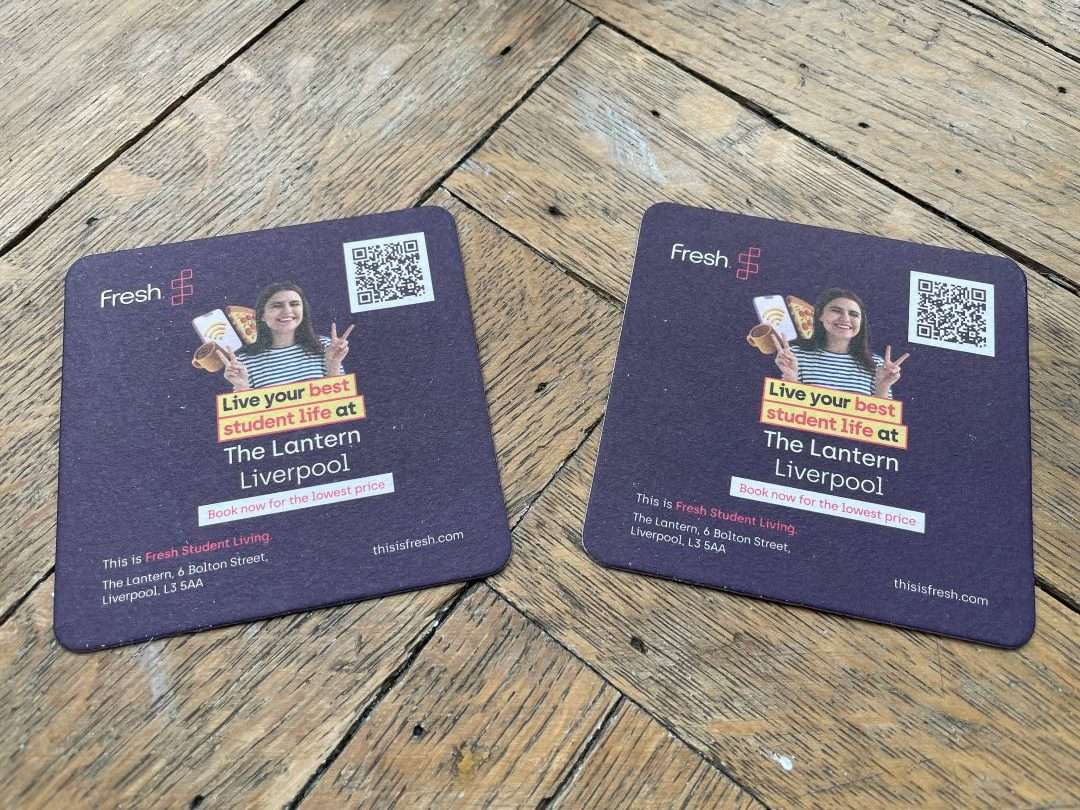 Two purple Fresh Student Living beer mats reading 'Live your best student life at The Lantern Liverpool' with a QR code, on a wooden table