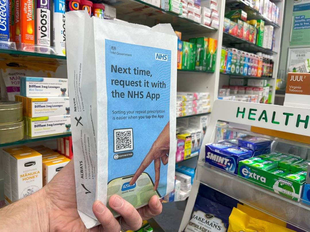 Hand holding a blue NHS England pharmacy bag reading 'Next time, request it with the NHS App' with a QR code, in a pharmacy