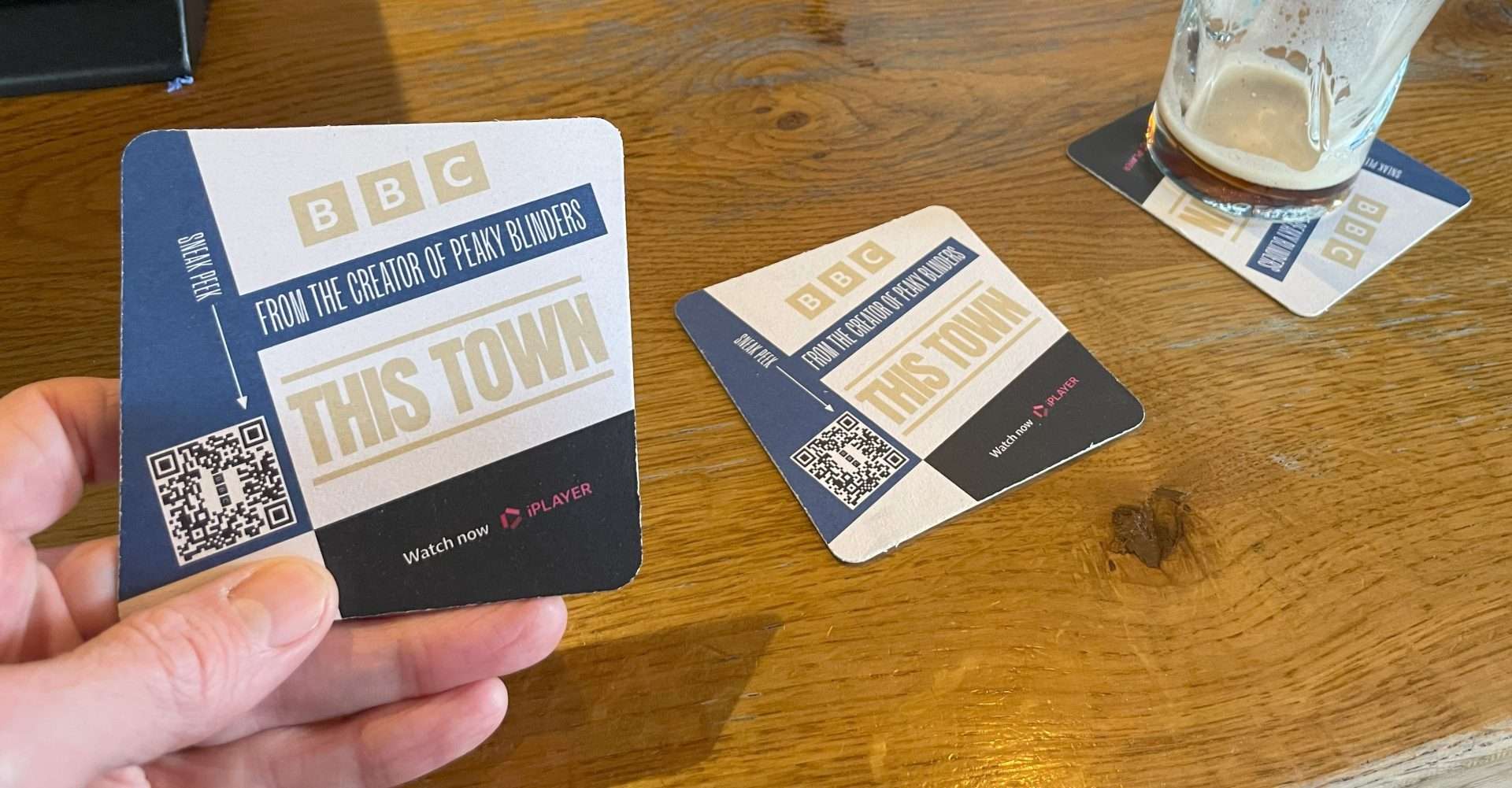 Hand holding a BBC This Town beer mat with a QR code to watch on iPlayer, with more mats and a pint glass on a pub table