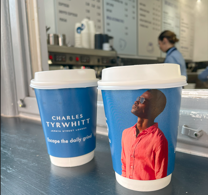 Two blue Charles Tyrwhitt takeaway coffee cups reading 'Escape the daily grind', featuring a man in a red shirt, on a coffee shop counter
