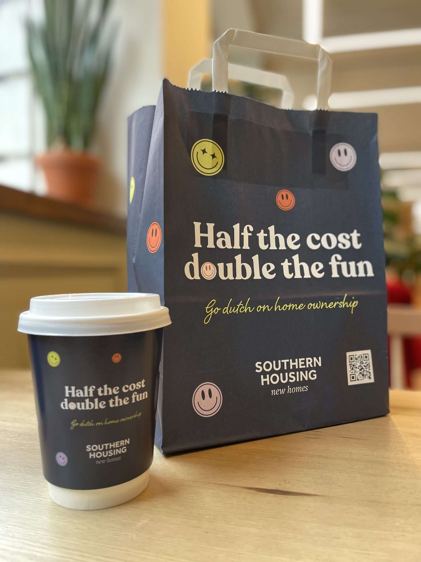 Navy Southern Housing coffee cup and deli bag reading 'Half the cost, double the fun - go dutch on home ownership', on a cafe table