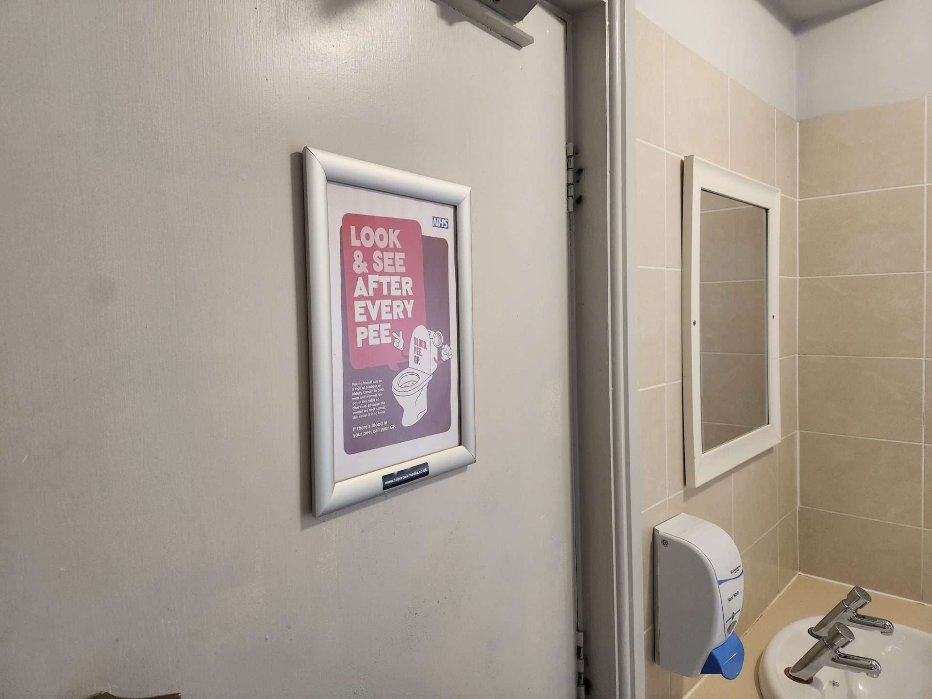Framed NHS Greater Manchester washroom poster reading 'Look & see after every pee' on a toilet door in a pub washroom