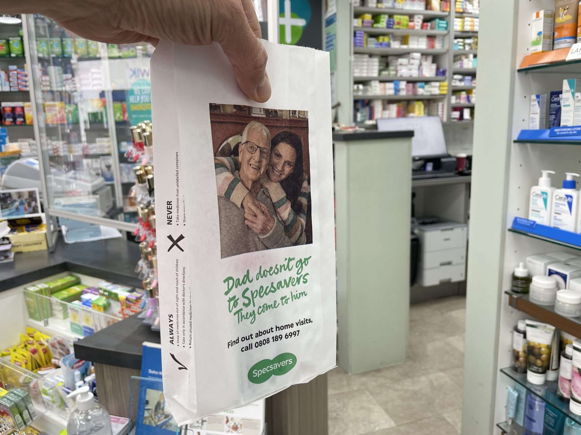 Hand holding a Specsavers pharmacy bag reading 'Dad doesn't go to Specsavers, they come to him' promoting home visits, in a pharmacy