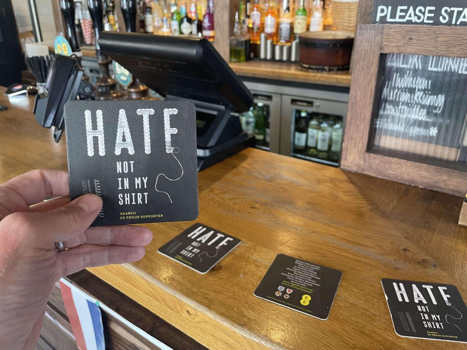 Hand holding a black EE 'Hate. Not in my shirt' beer mat at a pub bar, with more of the mats laid on the counter