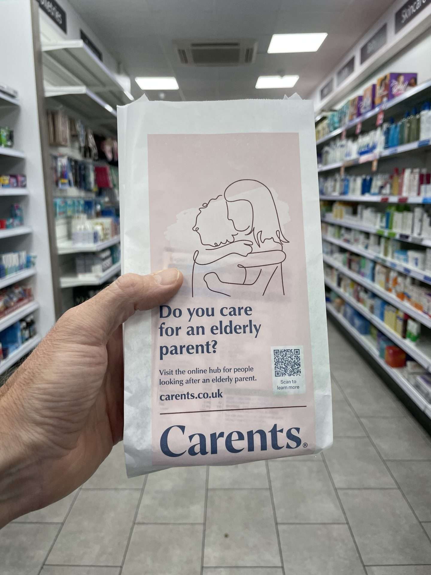 Hand holding a pink Carents pharmacy bag asking 'Do you care for an elderly parent?' with a QR code, in a pharmacy aisle