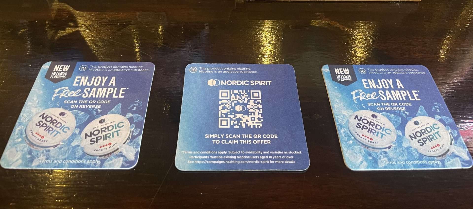 Three blue Nordic Spirit beer mats offering a free sample of new intense flavours, with a QR code to claim, on a dark pub table