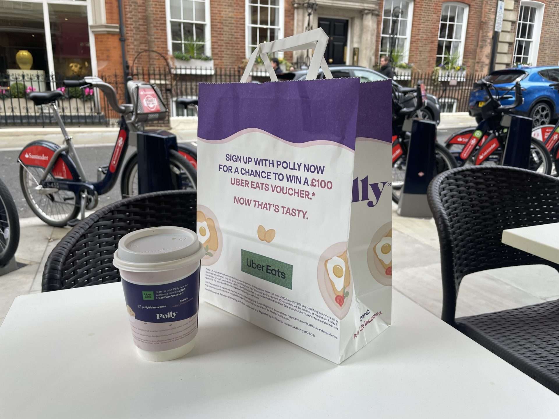 Polly life insurance coffee cup and takeaway bag offering a chance to win a 100 pound Uber Eats voucher, on an outdoor cafe table