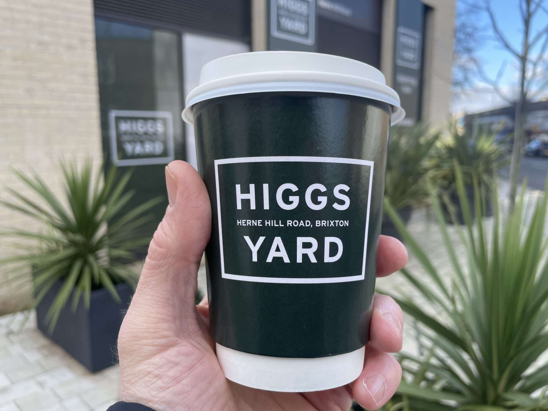 Hand holding a dark green Peabody Higgs Yard, Herne Hill Road, Brixton coffee cup outside the new development