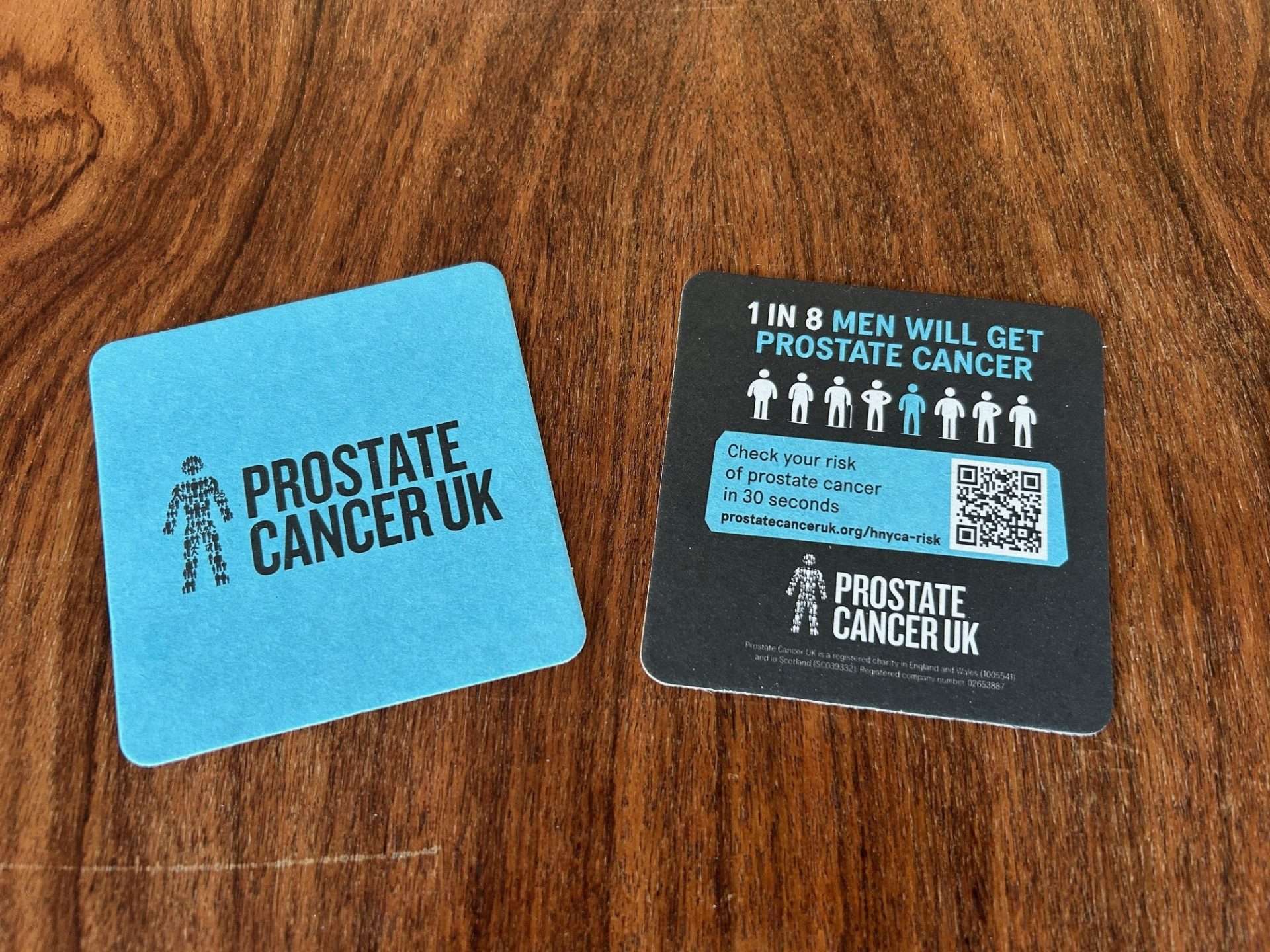 Two Prostate Cancer UK beer mats on a wooden table, one reading '1 in 8 men will get prostate cancer' with a QR code to check your risk