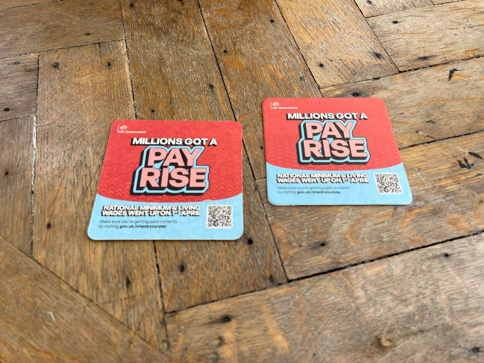 Two UK Government beer mats reading 'Millions got a pay rise' about the National Minimum and Living Wage increase, on a wooden floor