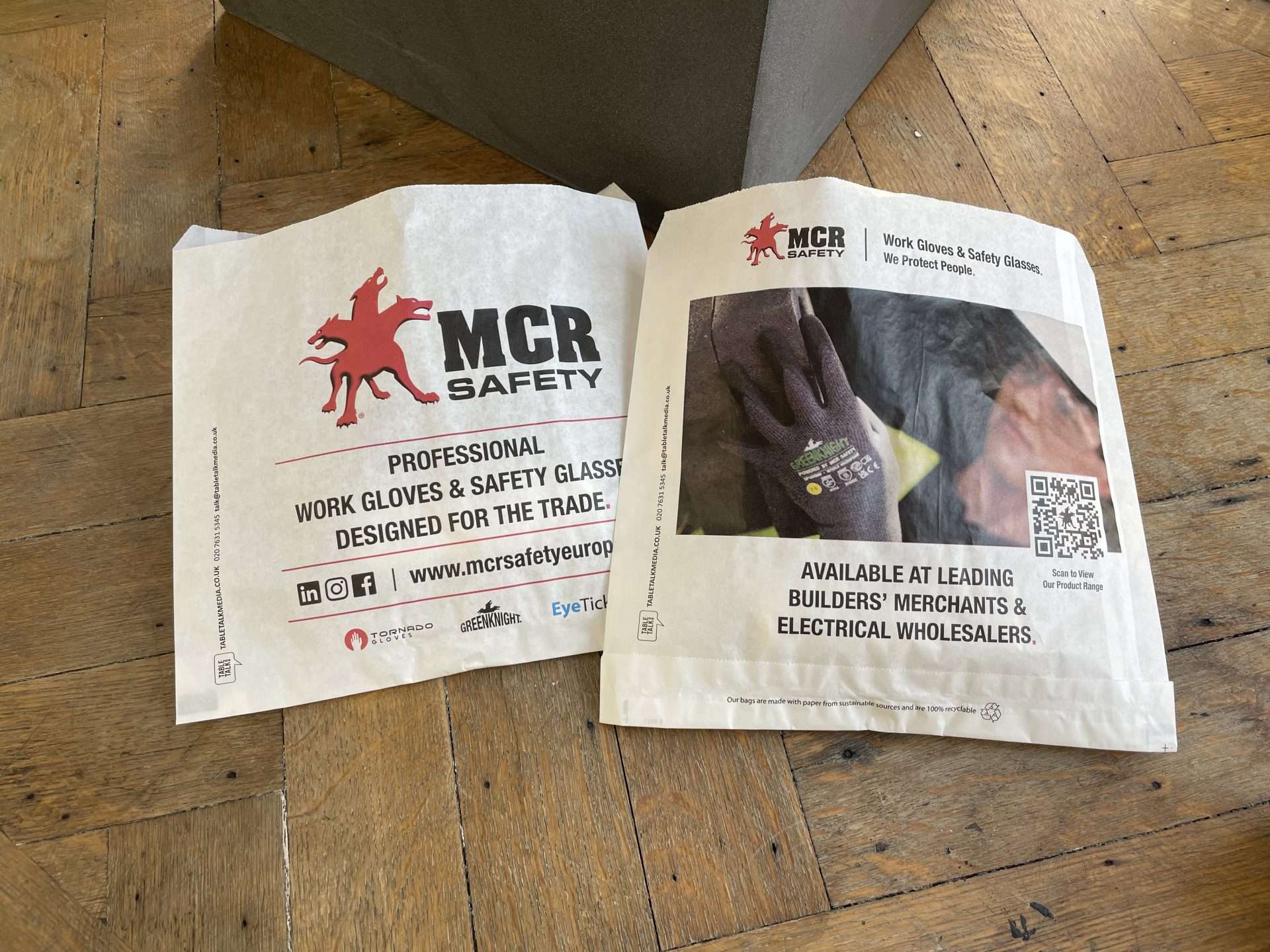 Two MCR Safety bacon butty bags advertising professional work gloves and safety glasses for the trade, on a wooden floor