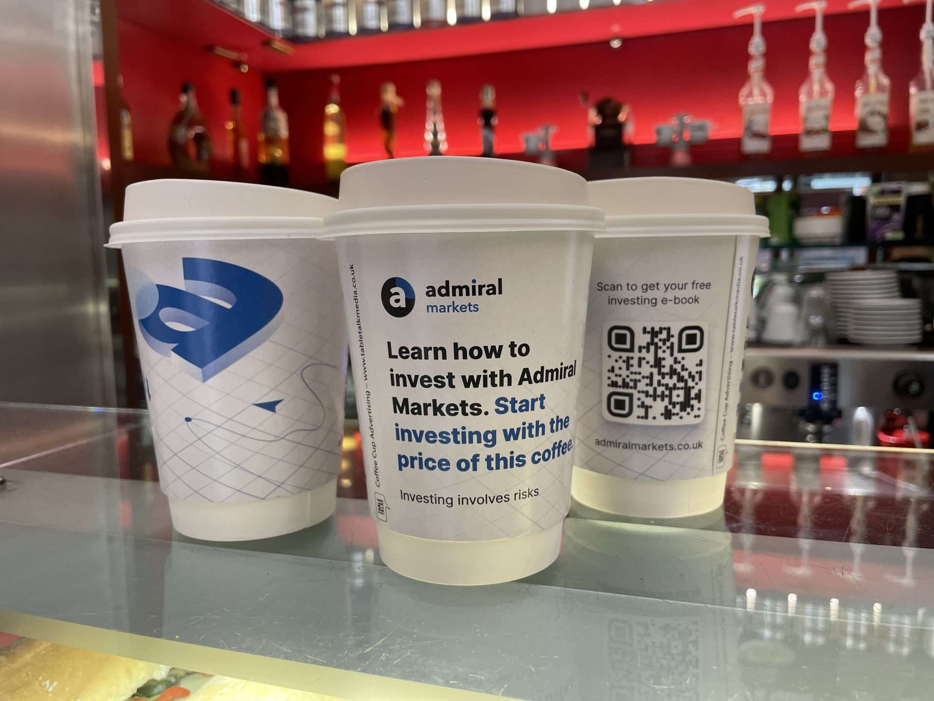 Three Admiral Markets coffee cups reading 'Start investing with the price of this coffee' with a QR code for a free e-book, on a cafe counter
