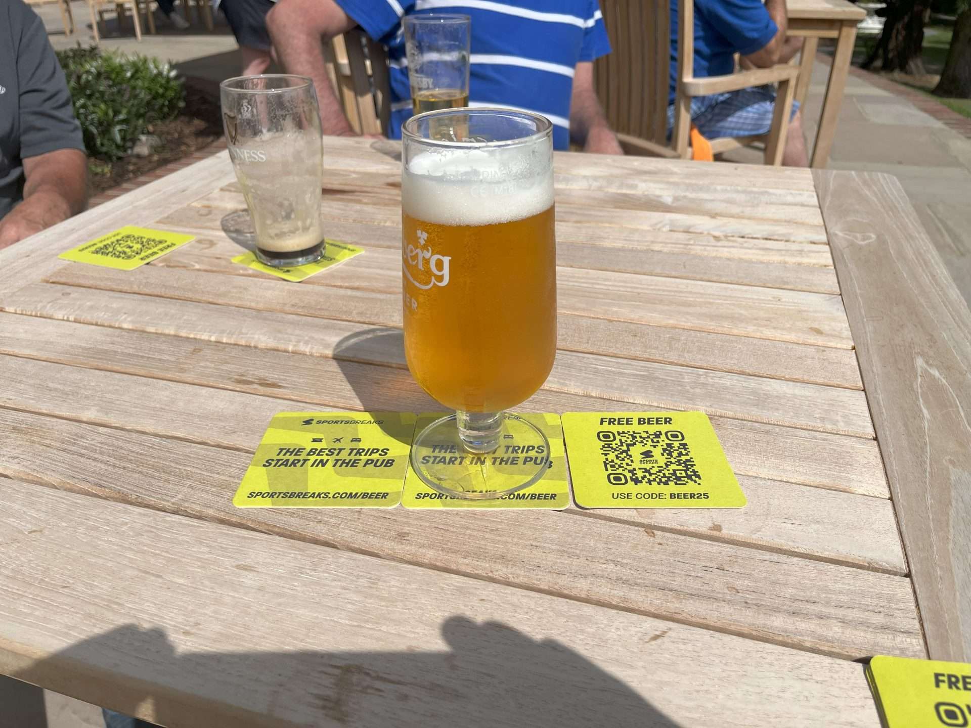 Pint of beer on yellow Sportsbreaks.com beer mats reading 'The best trips start in the pub' with a free beer QR code, on a pub garden table