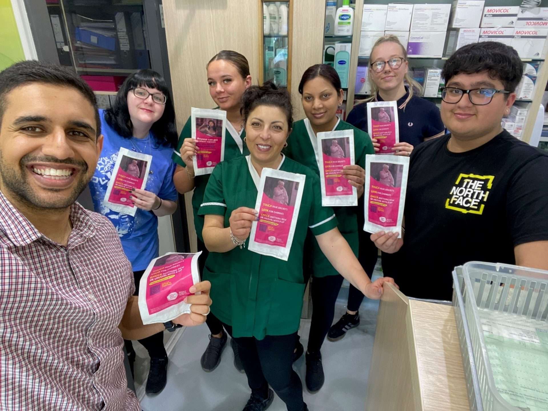 Smiling pharmacy team holding up pink Breast Cancer Now Touch, Look, Check pharmacy bags inside their pharmacy