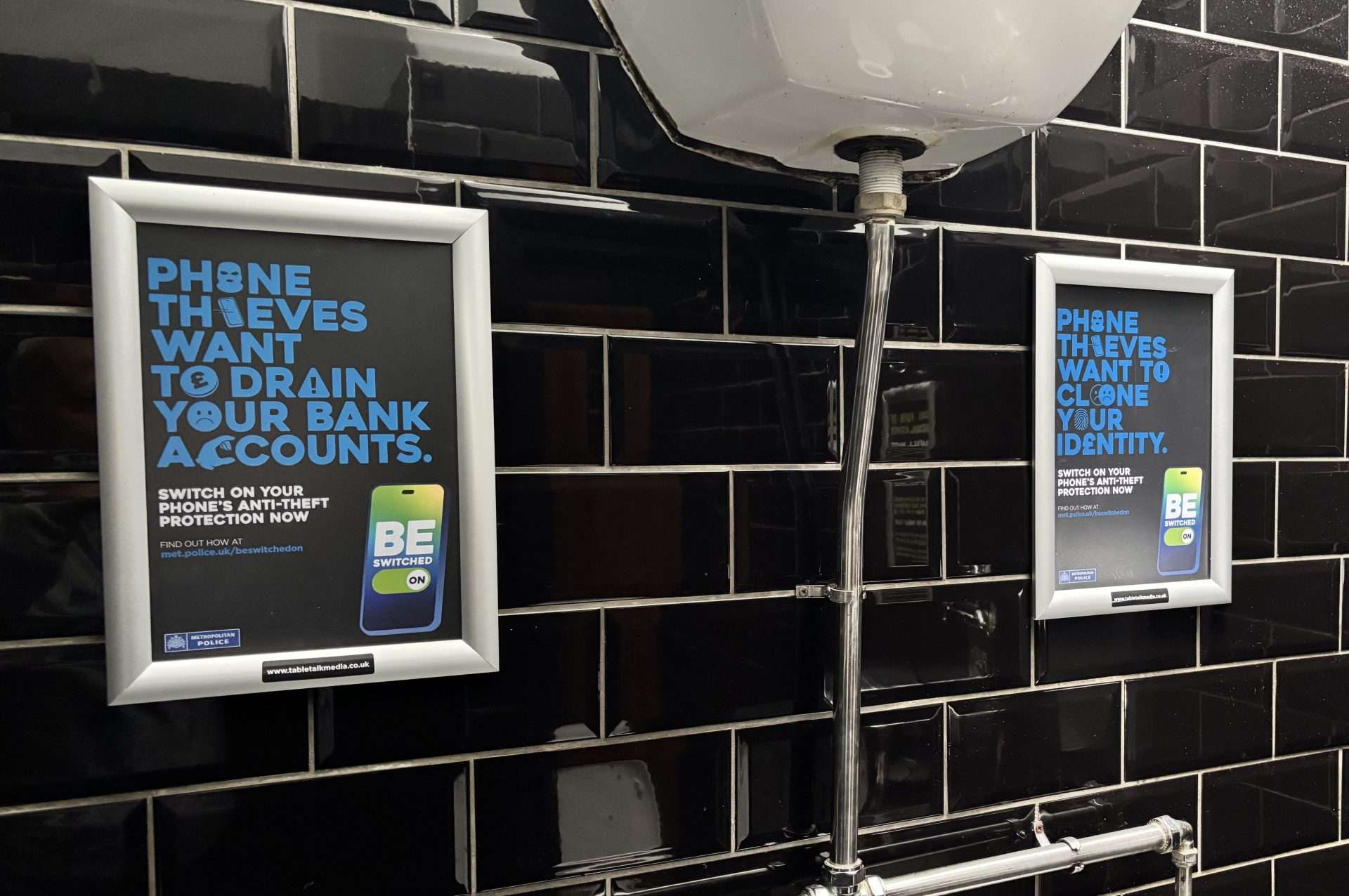 Two framed Met Police 'Be Switched On' posters warning about phone thieves, on a black tiled pub washroom wall