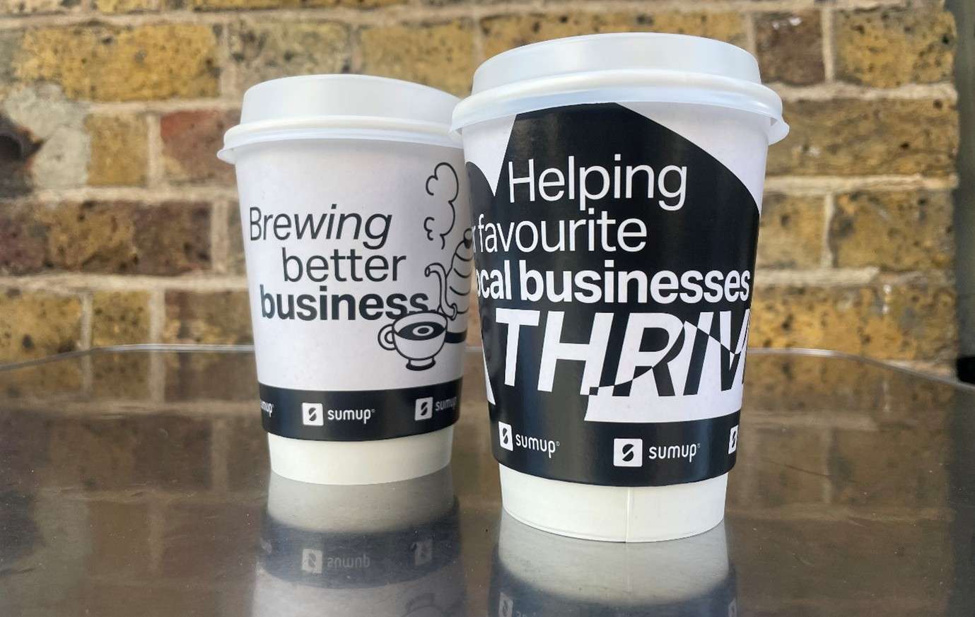 Two black and white SumUp takeaway coffee cups reading 'Brewing better business' and 'Helping your favourite local businesses thrive', against a brick wall