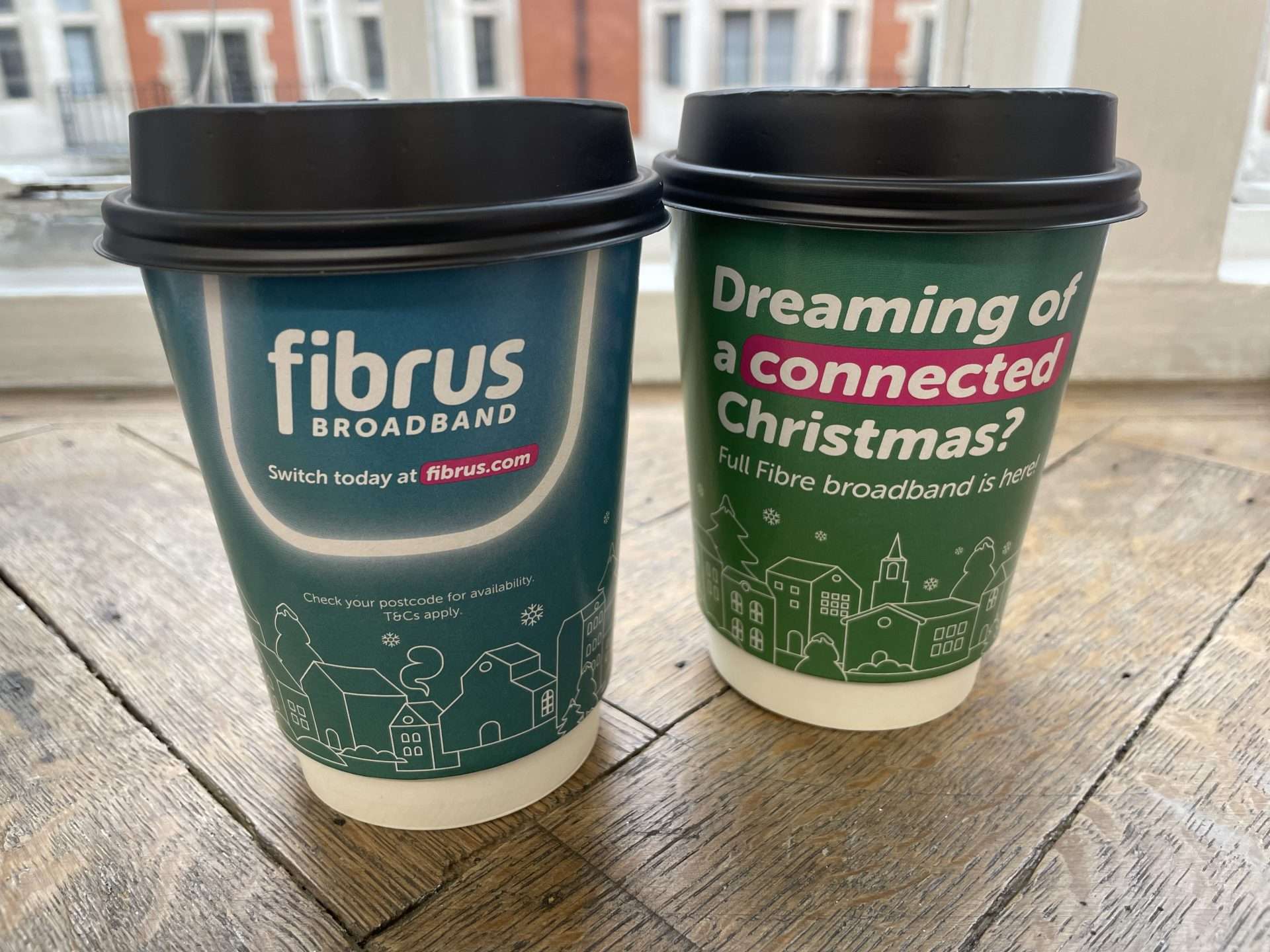 Two Fibrus Broadband takeaway coffee cups, one reading 'Dreaming of a connected Christmas?' with a festive town illustration