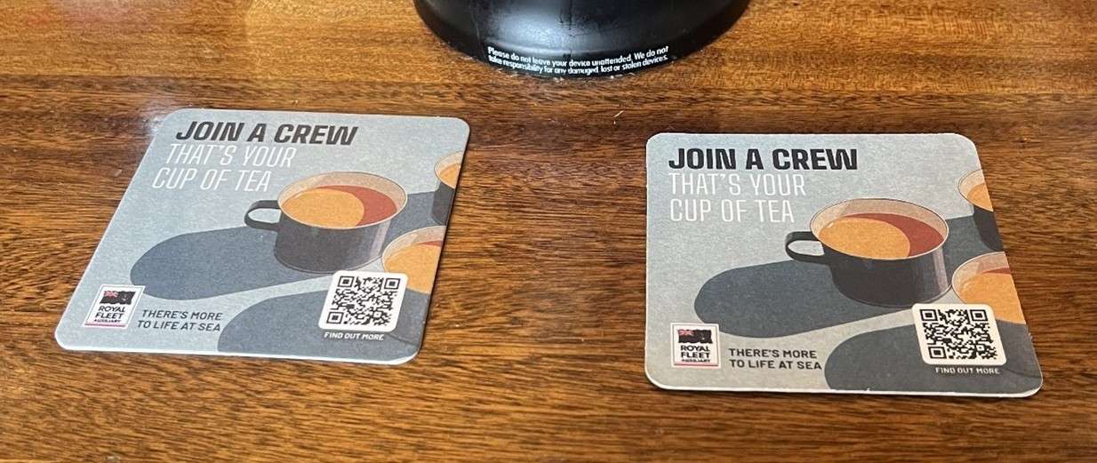Two Royal Fleet Auxiliary beer mats reading 'Join a crew that's your cup of tea' with a QR code, on a wooden pub table