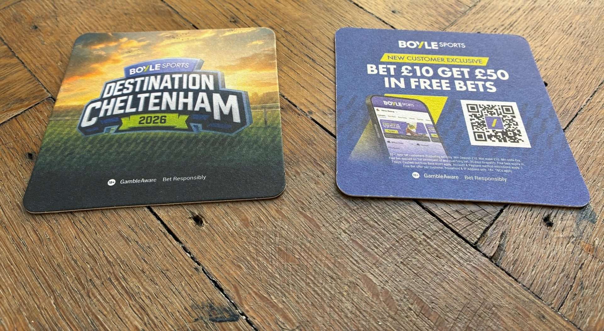 Two BoyleSports beer mats on a wooden table, one reading 'Destination Cheltenham 2026' and one with a new customer free bets offer and QR code