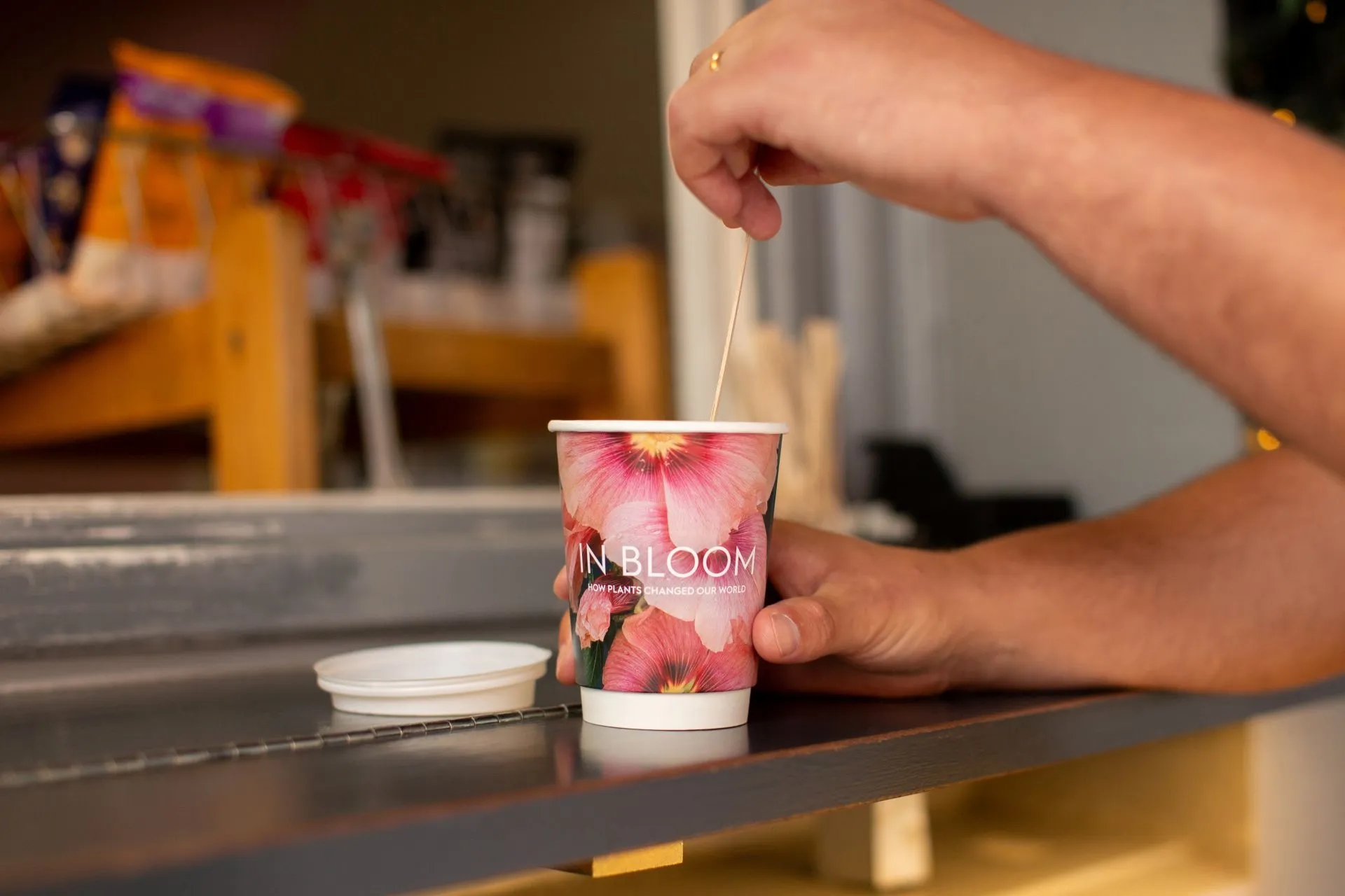 Customer stirring an Ashmolean In Bloom floral takeaway coffee cup at a café counter