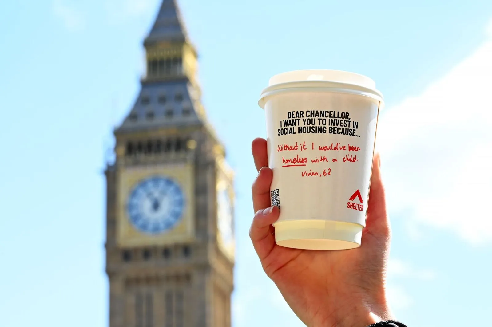 Hand holding a Shelter 'Dear Chancellor' social housing coffee cup with a handwritten message in front of Big Ben
