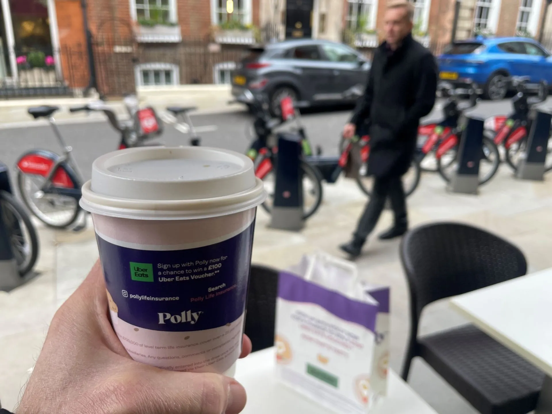 Hand holding a Polly life insurance-branded takeaway coffee cup at a London street café