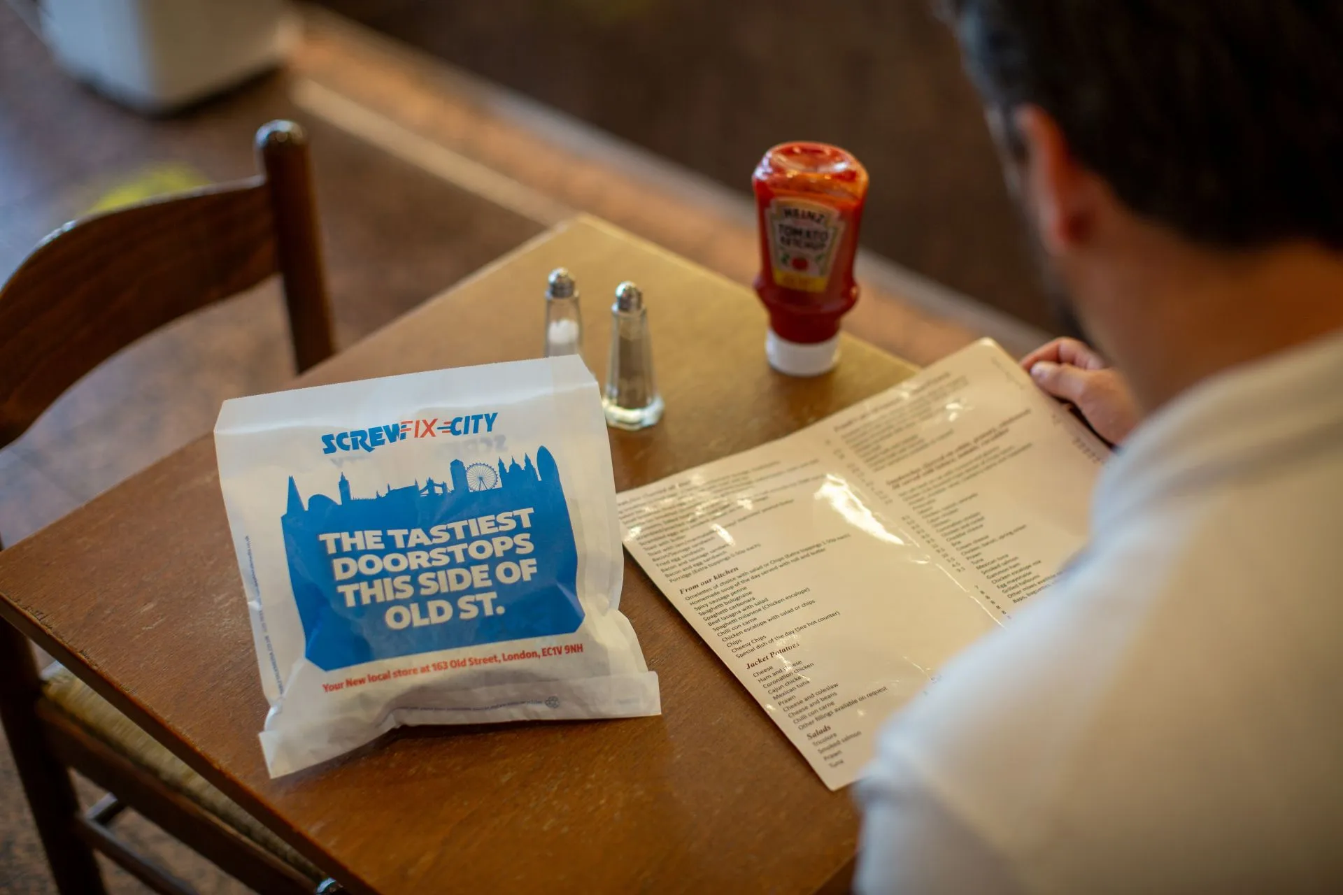 Screwfix City 'The tastiest doorstops this side of Old St.' sandwich bag on a café table beside a menu