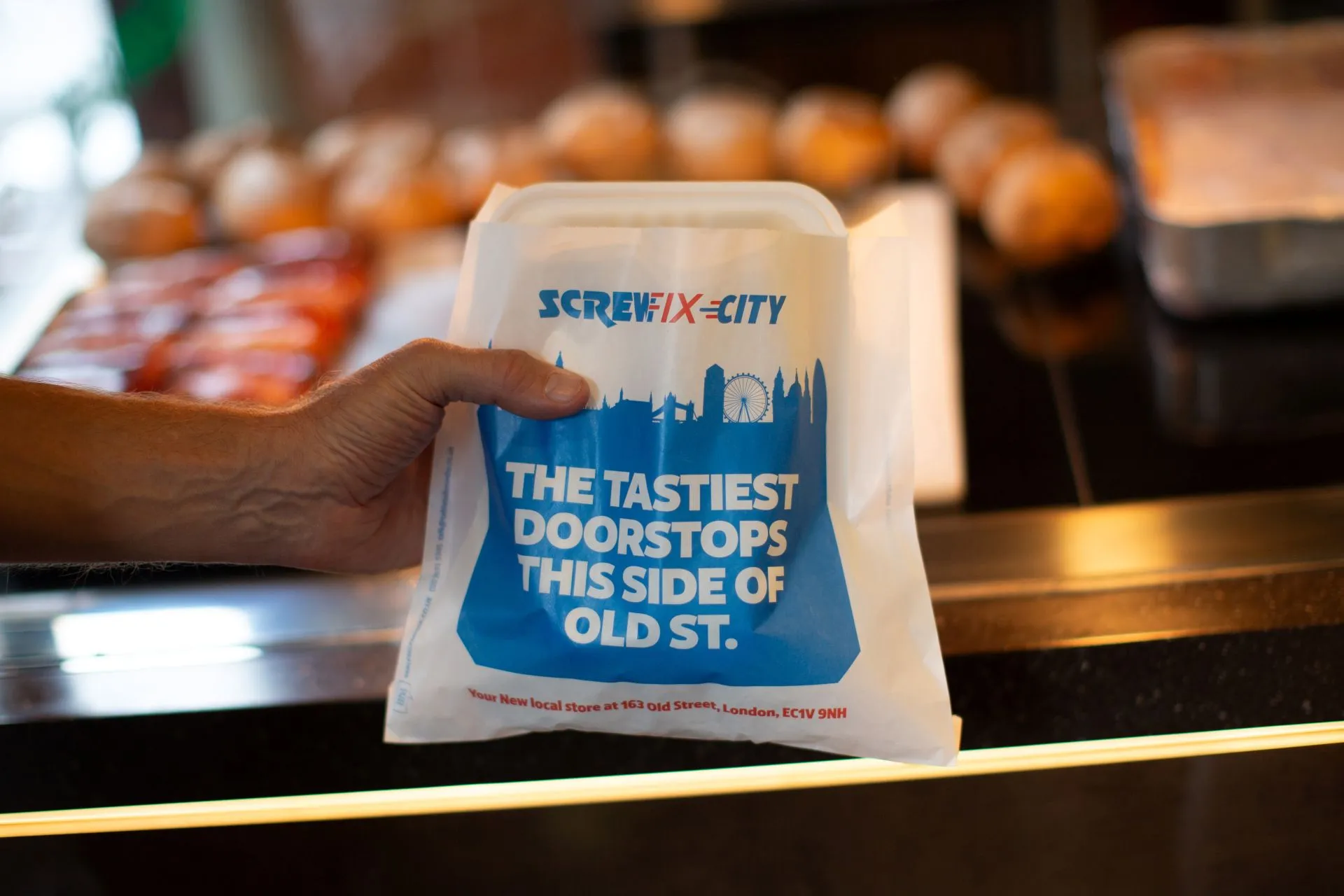 Hand holding a Screwfix City 'The tastiest doorstops this side of Old St.' sandwich bag over a bakery counter