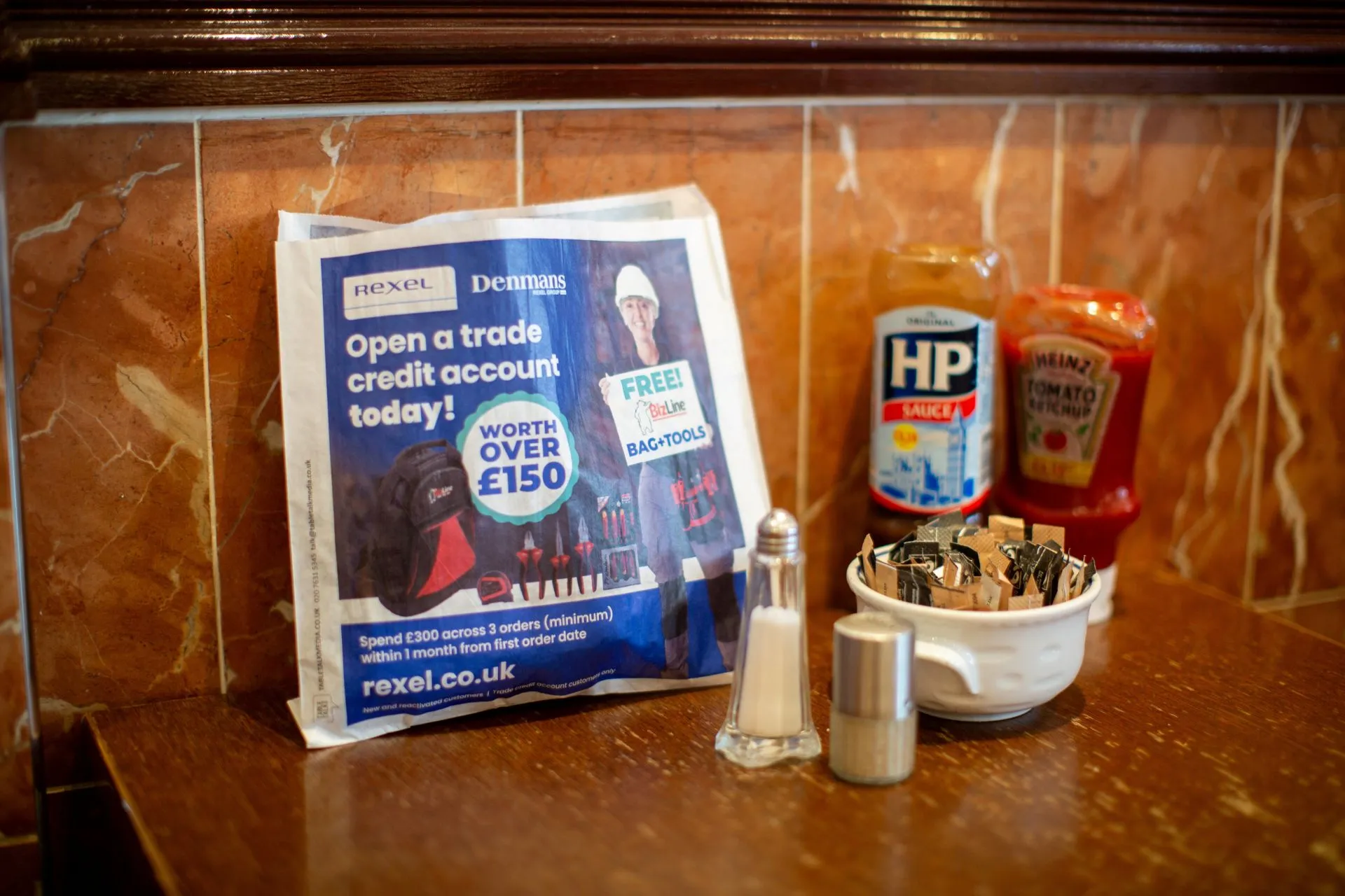 Rexel and Denmans trade credit account sandwich bag on a café table beside HP Sauce and condiments