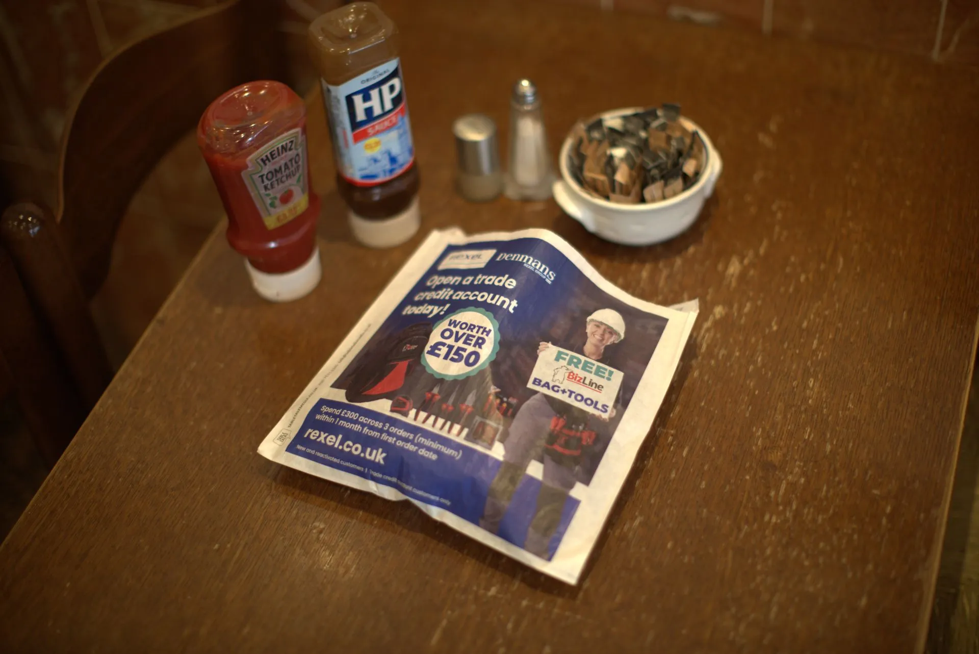 Rexel and Denmans trade credit sandwich bag on a wooden café table next to ketchup, HP Sauce and sugar sachets