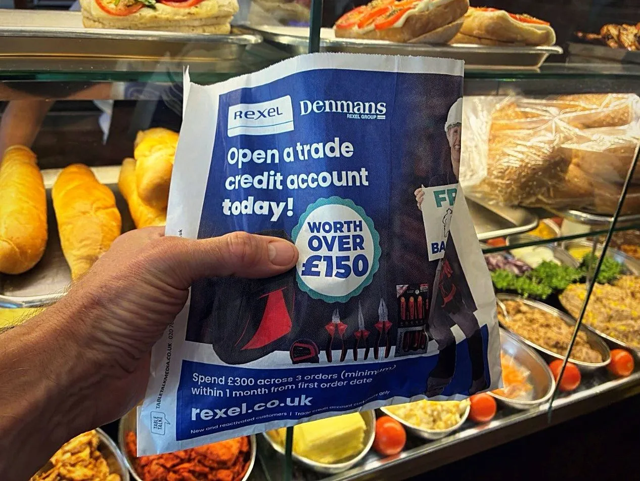 Hand holding a Rexel and Denmans 'Open a trade credit account' sandwich bag in front of a sandwich bar counter
