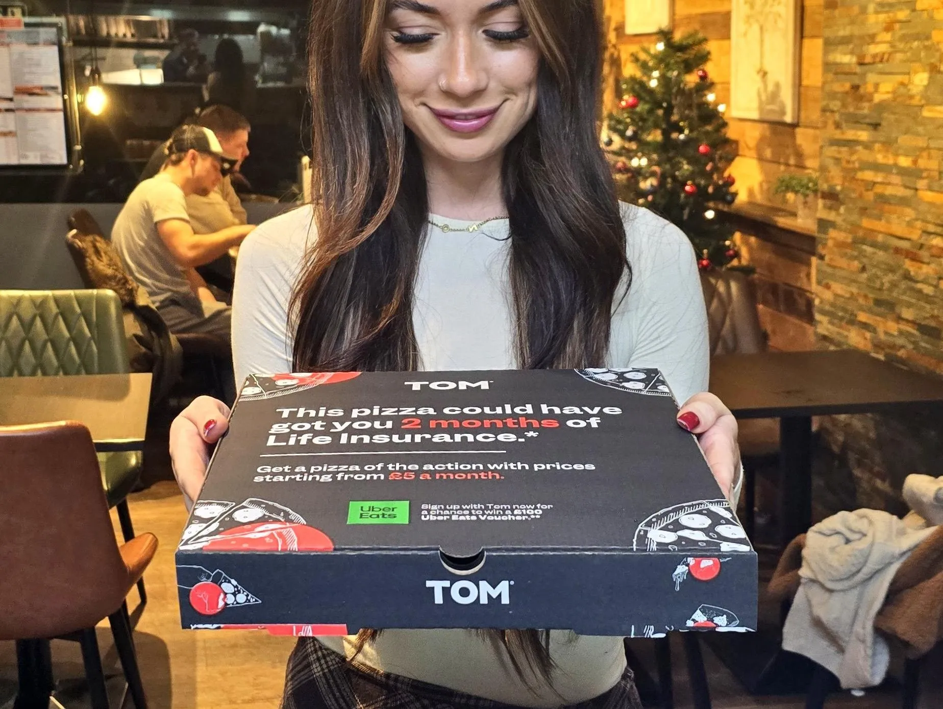 Woman holding a TOM Life Insurance 'This pizza could have got you 2 months of life insurance' pizza box in a restaurant