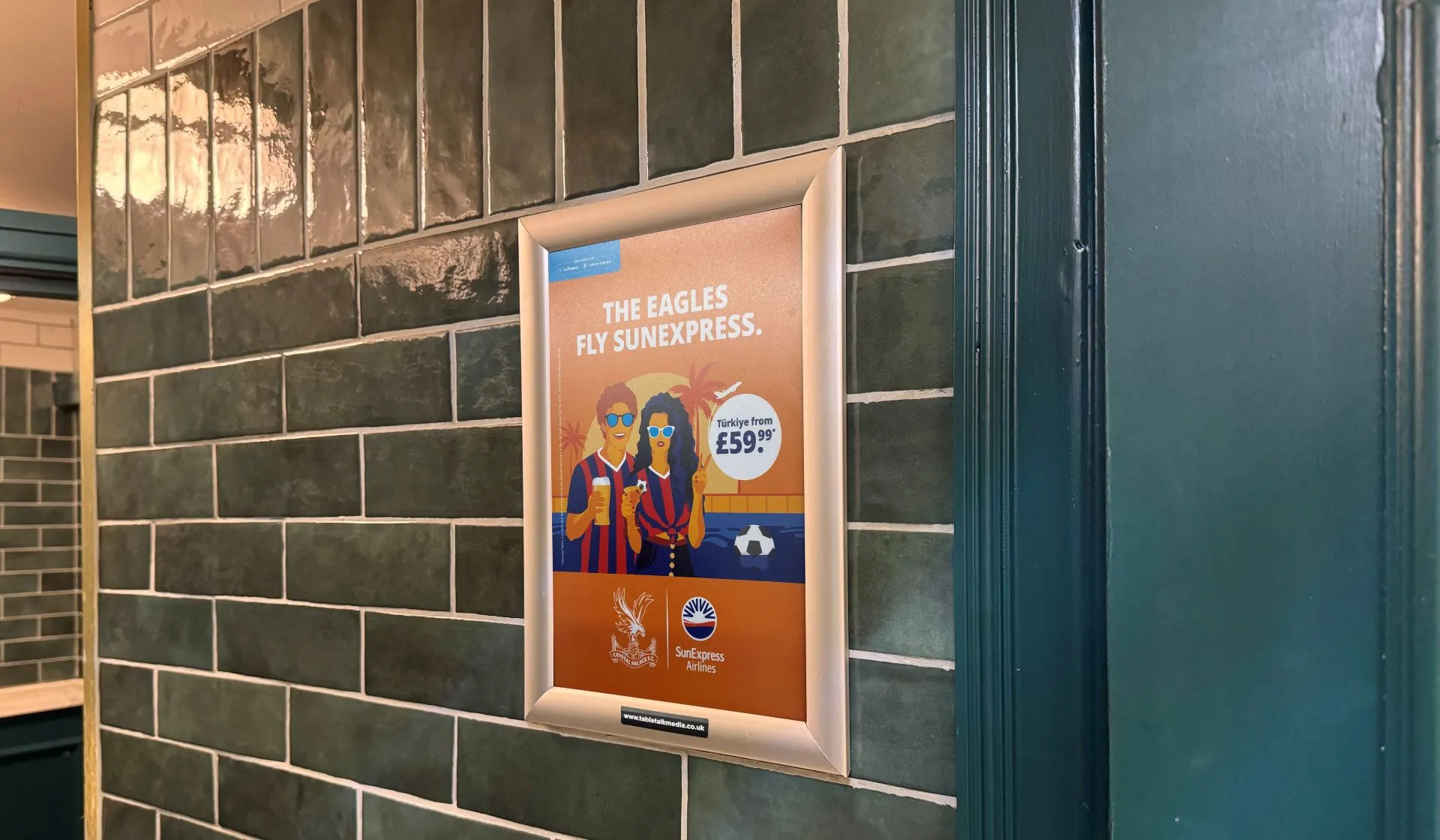 SunExpress Crystal Palace poster in a framed washroom panel on a green-tiled pub washroom wall