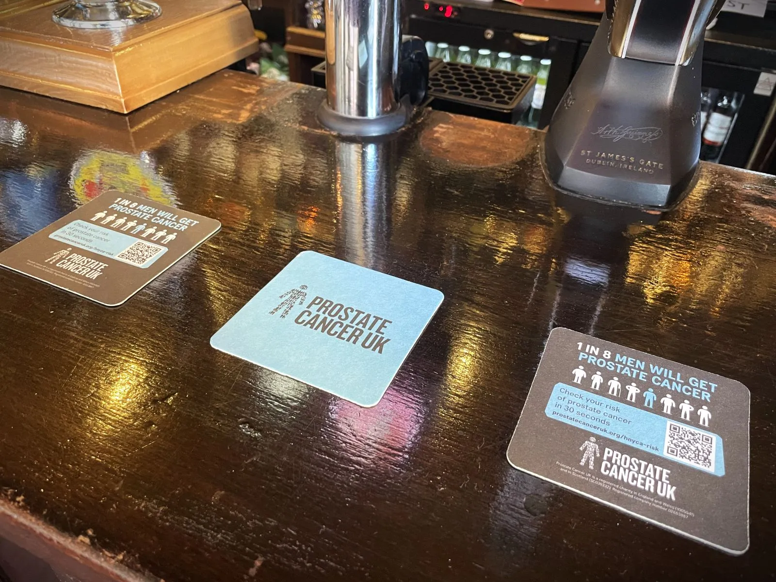 Three Prostate Cancer UK beer mats with '1 in 8 men' message and QR code on a pub bar top