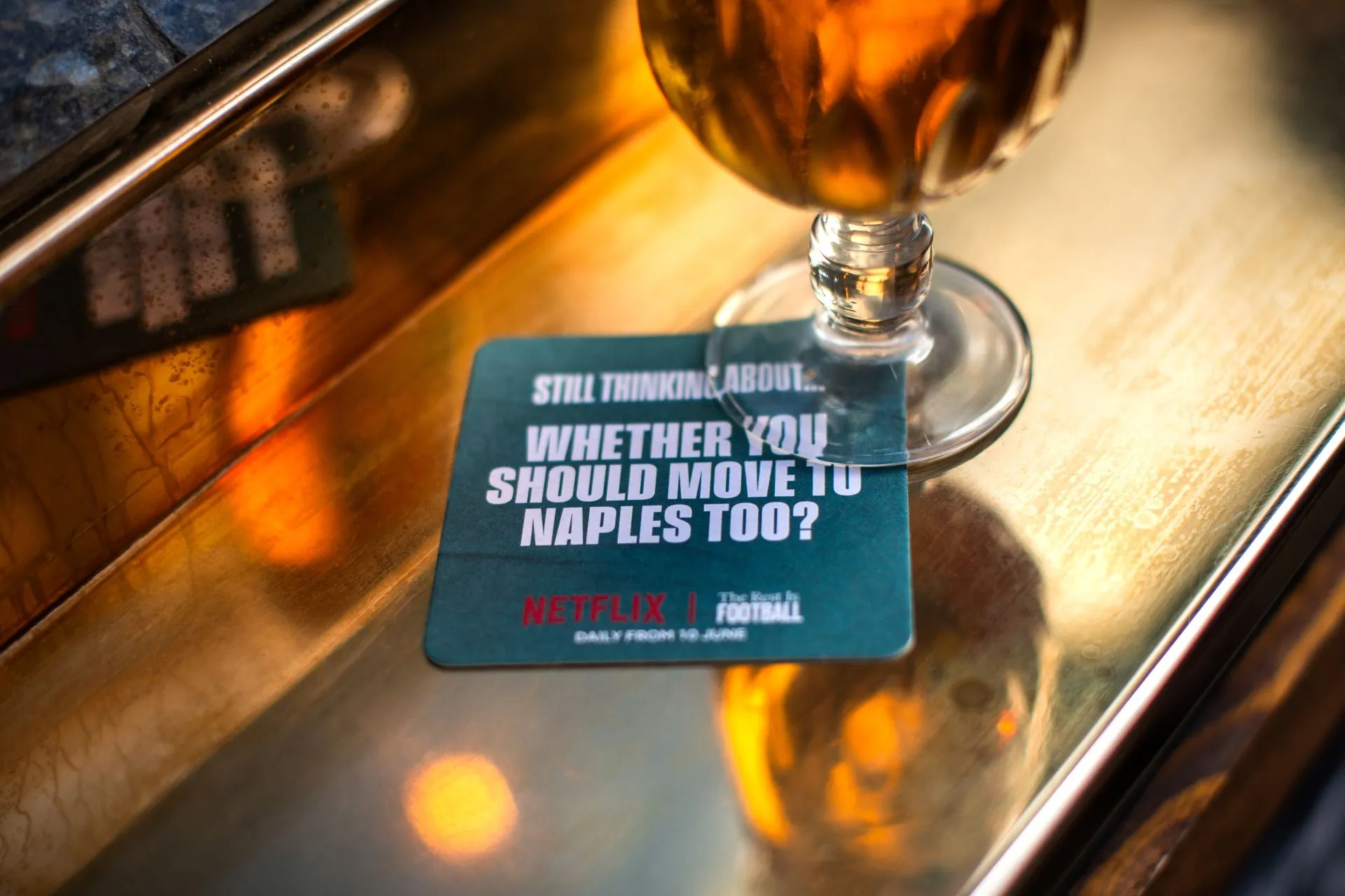 Netflix The Rest Is Football beer mat under a glass of beer on a brass pub bar