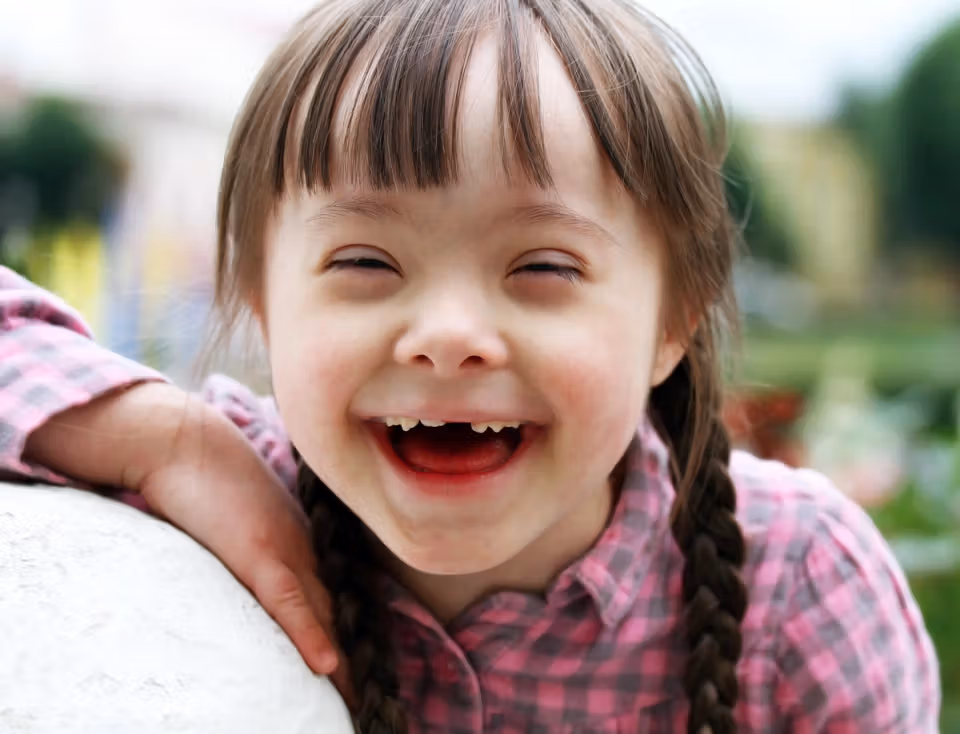 Smiling young girl with braided hair wearing a pink checkered shirt outdoors, leaning on a white surface.