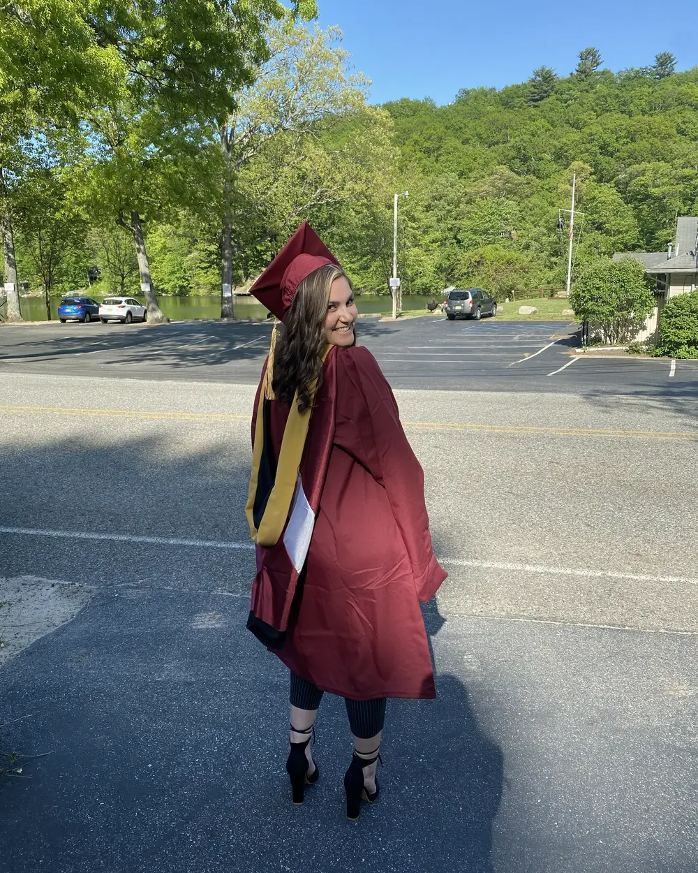 Amanda in a maroon graduation gown and cap, looking back over her shoulder on a tree-lined road