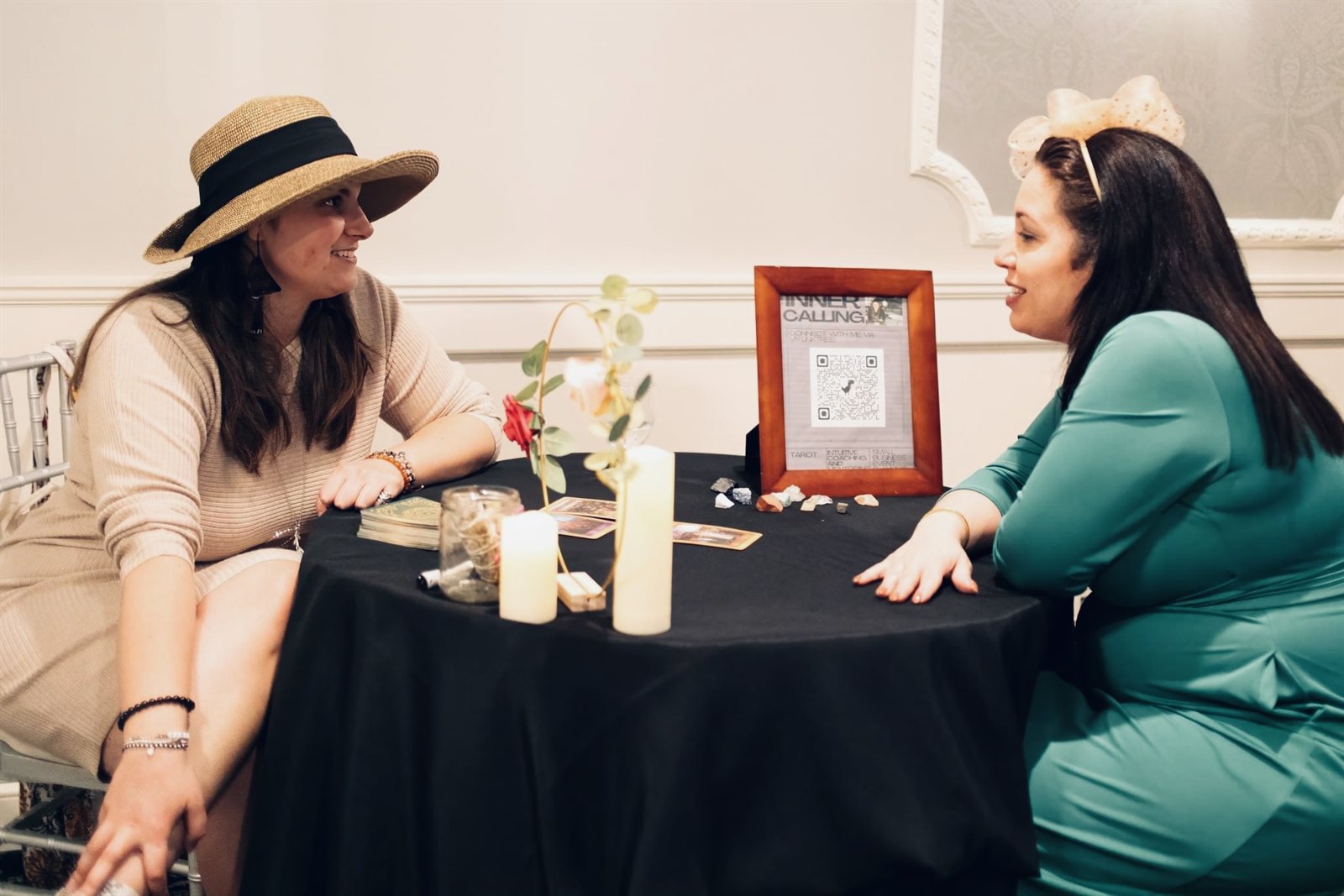 Amanda in a straw hat leaning in to talk with a client across a small black table, a framed QR code and candles between them