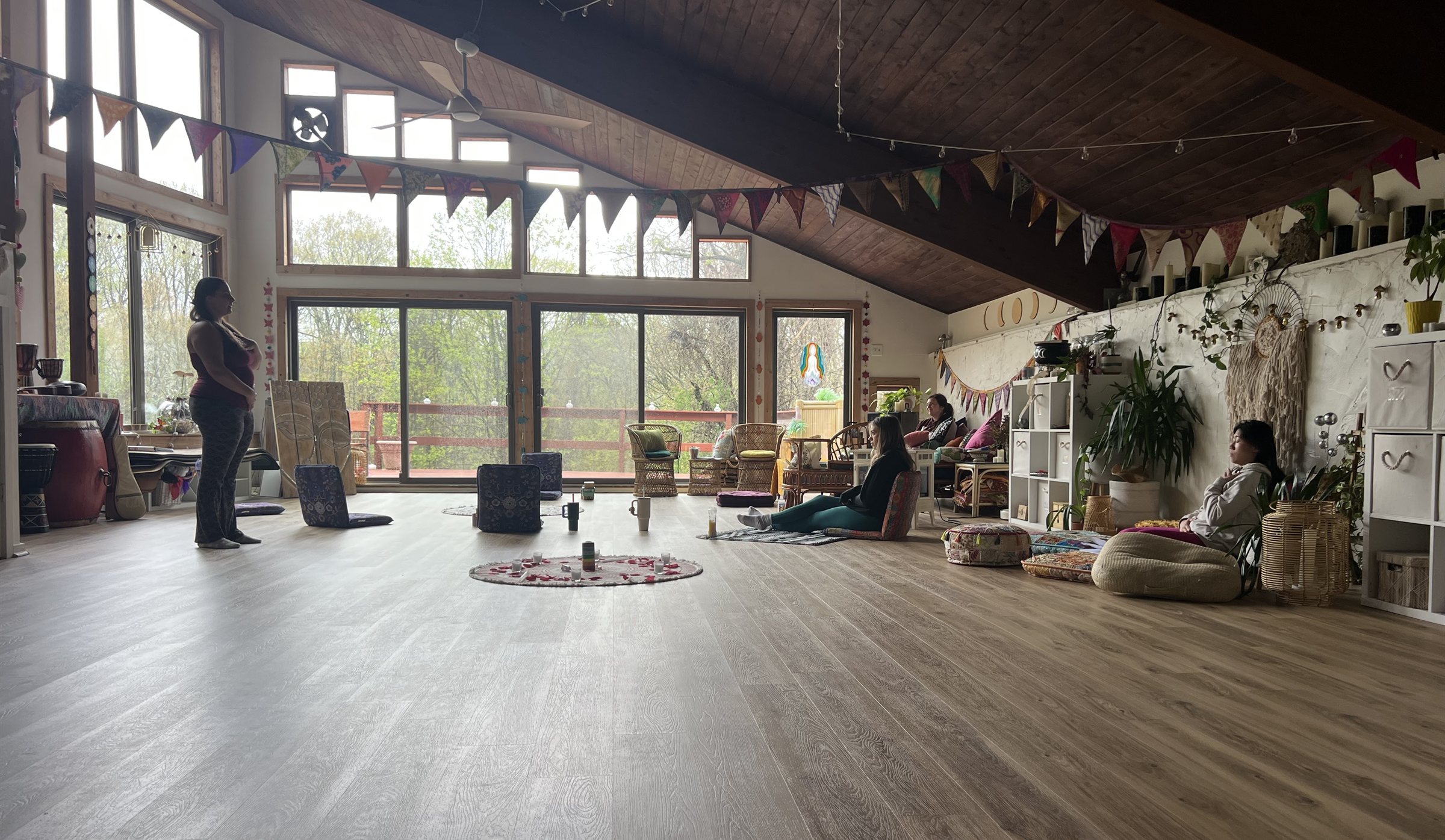 A bright studio with a high wooden ceiling and strings of flags, a small circle of flowers on the floor and a few people sitting and standing around the room
