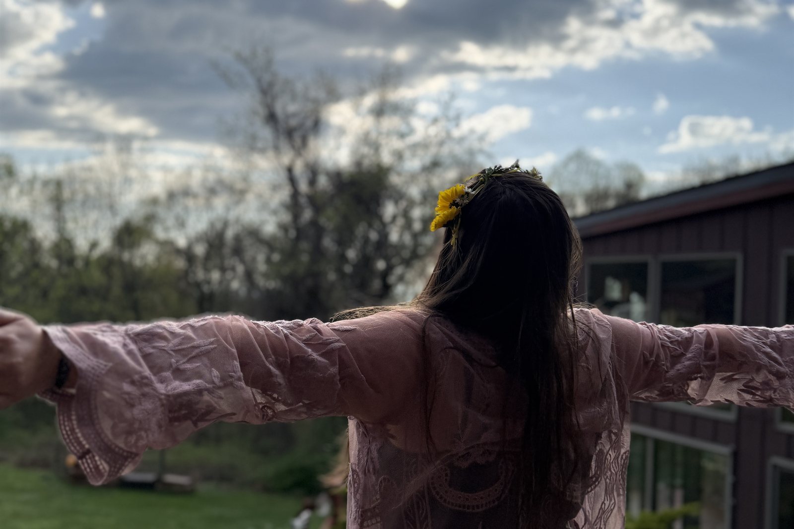 Amanda seen from behind with her arms stretched wide toward a cloudy sky, a yellow flower in her hair