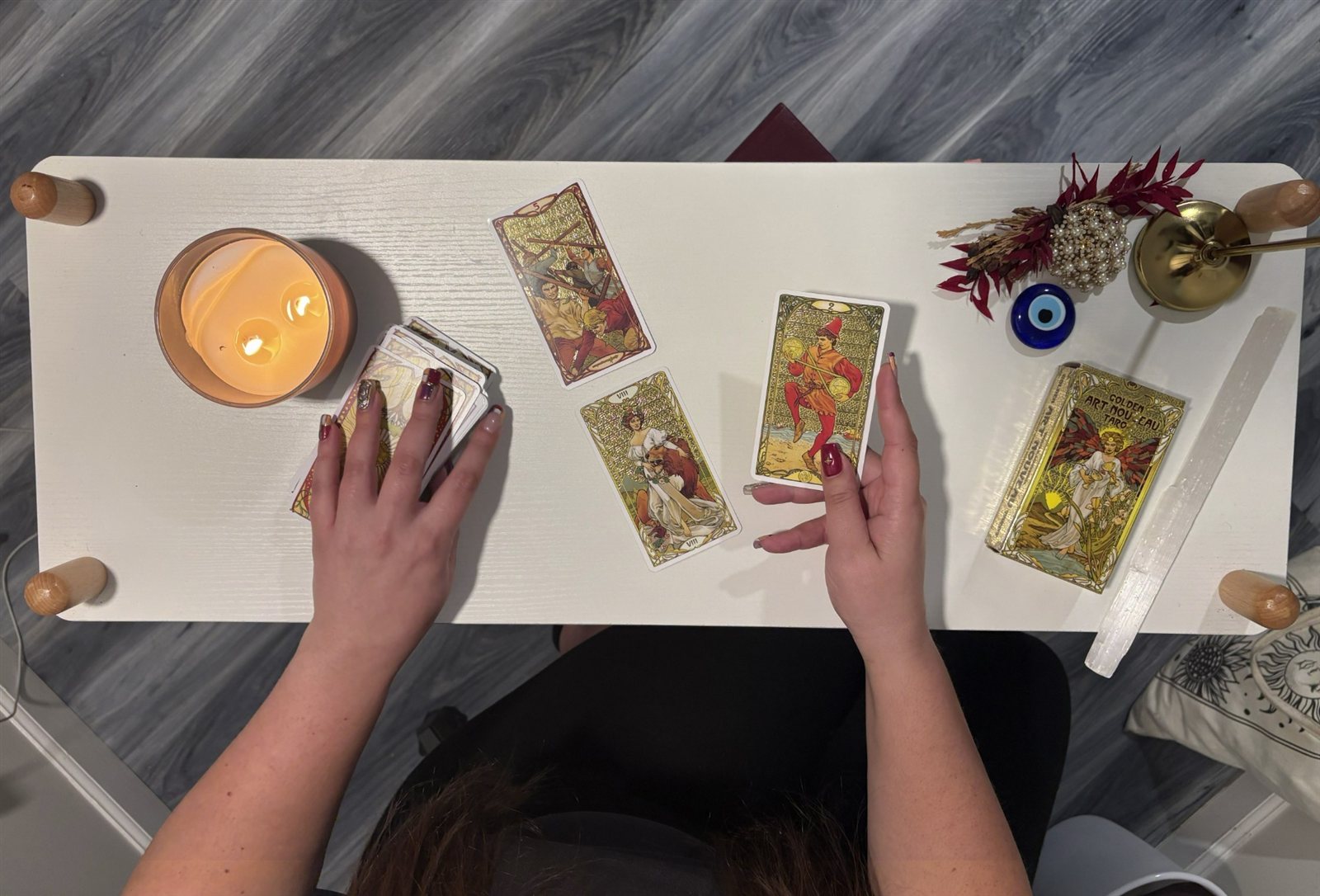 Looking down on a white reading table: Amanda holding one tarot card while her other hand rests on the deck, three cards laid out, a lit candle, a tarot box, a selenite wand and an evil eye charm