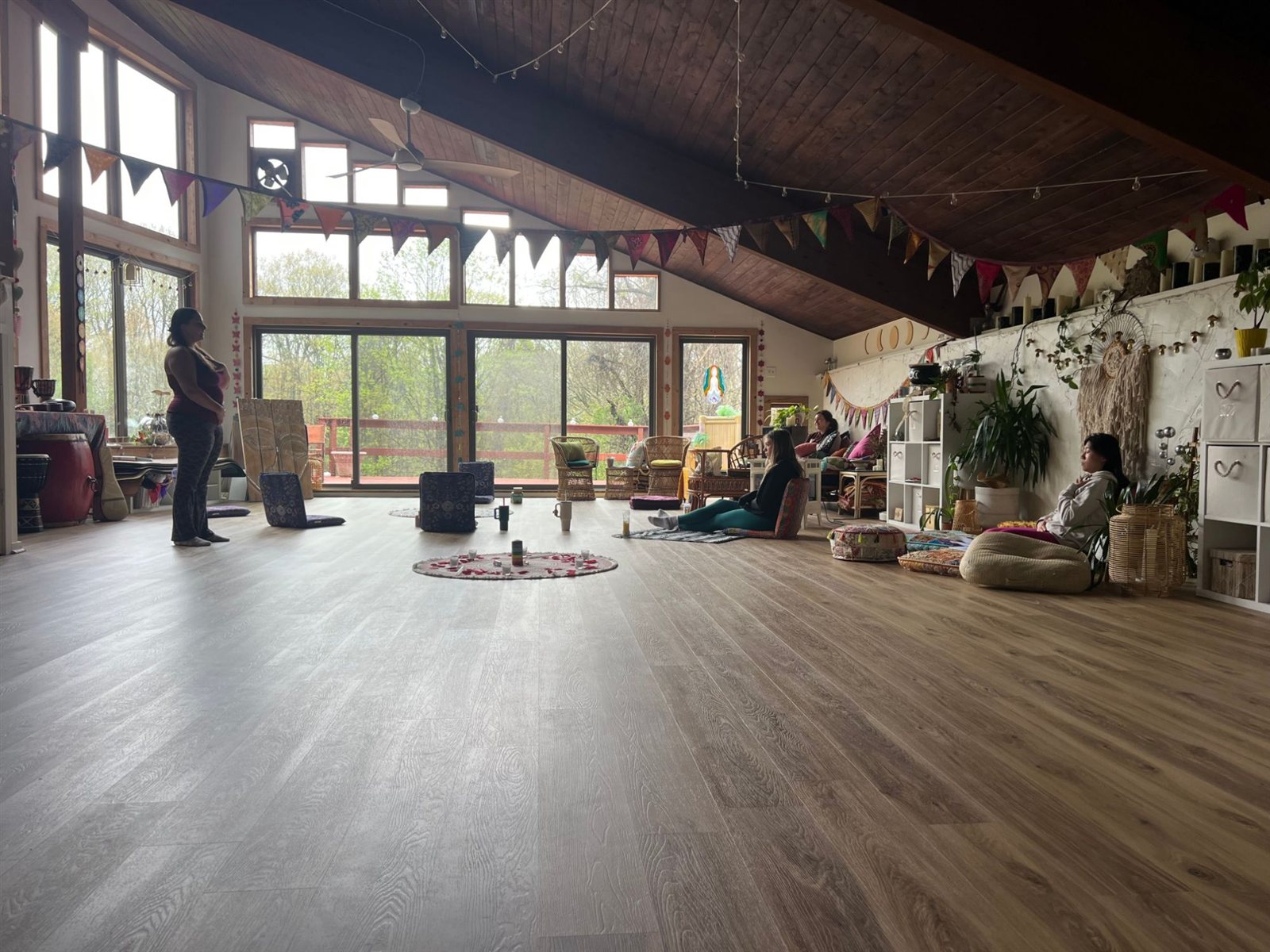 A bright studio with a high wooden ceiling and strings of flags, a small circle of flowers and candles on the floor, and a few people sitting and standing around the room