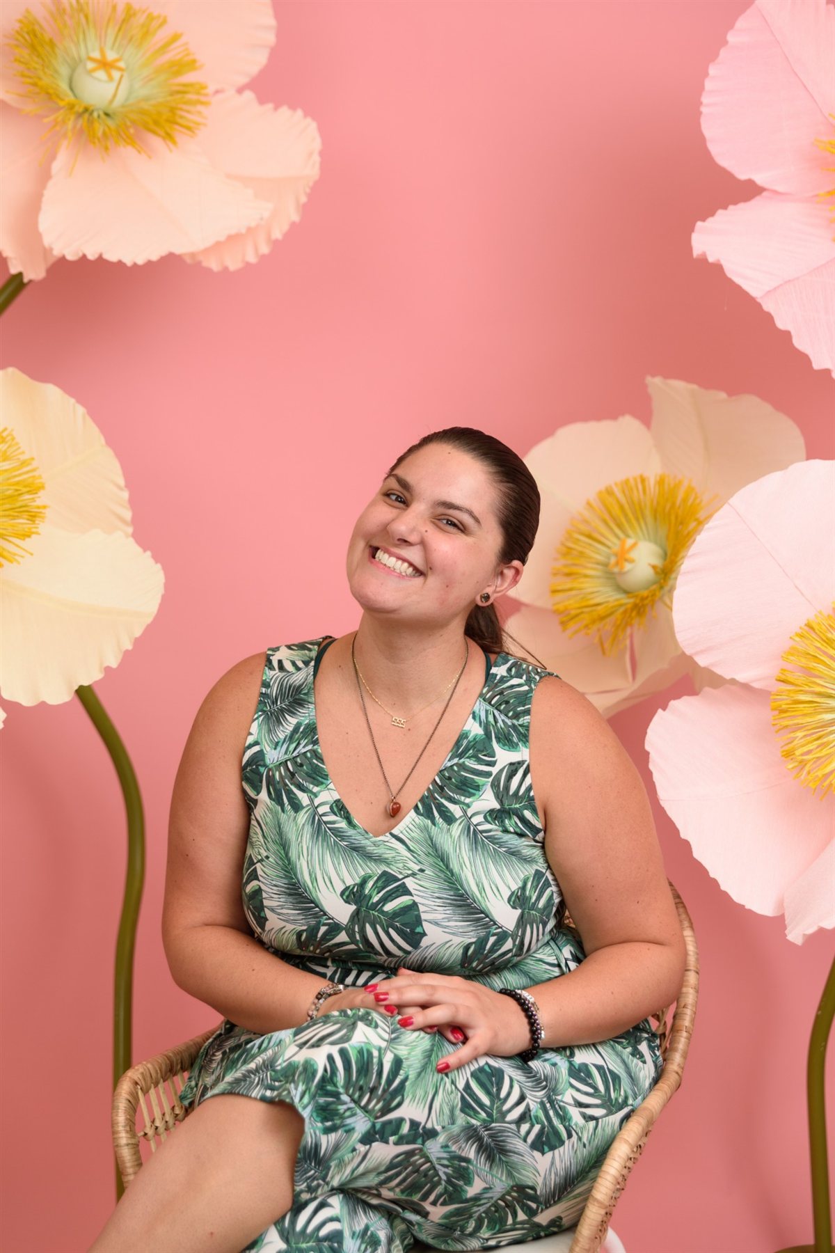 Amanda smiling with her head tilted, seated in a wicker chair in a leaf print dress against a pink backdrop of oversized paper poppies