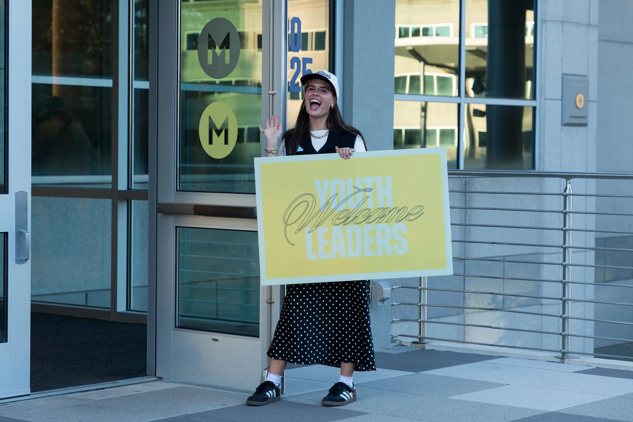 Young woman standing outside a building entrance, smiling and waving while holding a large yellow sign that says 'Welcome Youth Leaders.' She is dressed in a black and white polka dot skirt, sneakers, and a cap.