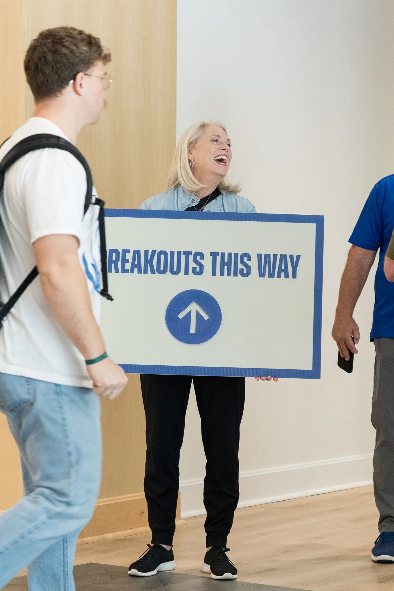 A smiling woman holding a large sign that says 'BREAKOUTS THIS WAY' with an upward arrow, standing indoors near a wooden wall, while two people walk nearby.