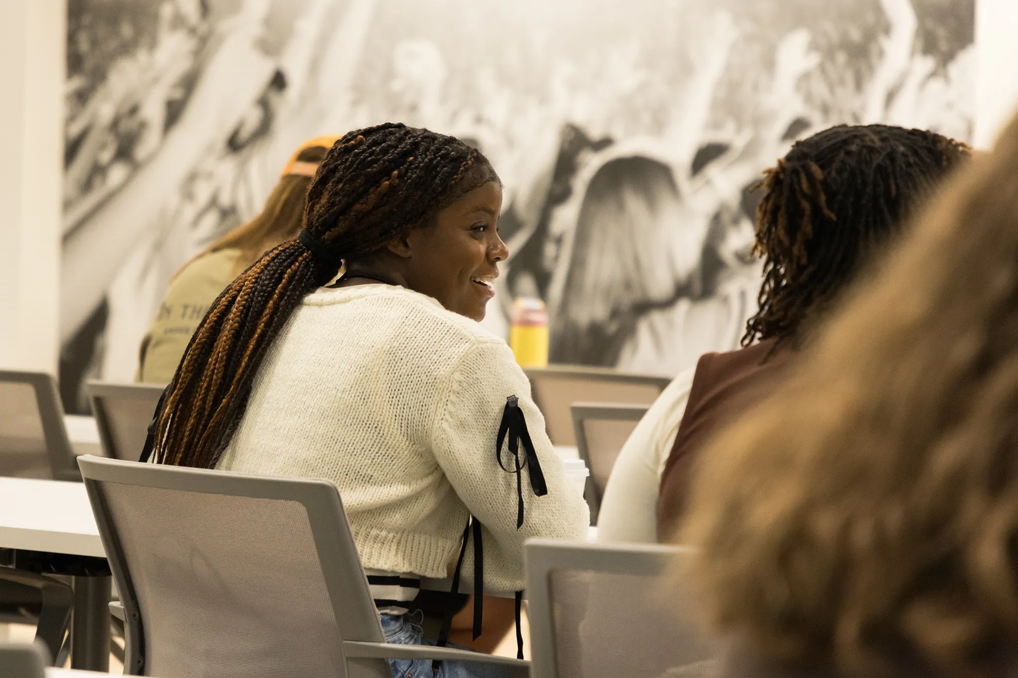Young woman with long braids tied back, wearing a white sweater with black ribbon details on the sleeves, sitting in a classroom or conference room, smiling and talking with another person.