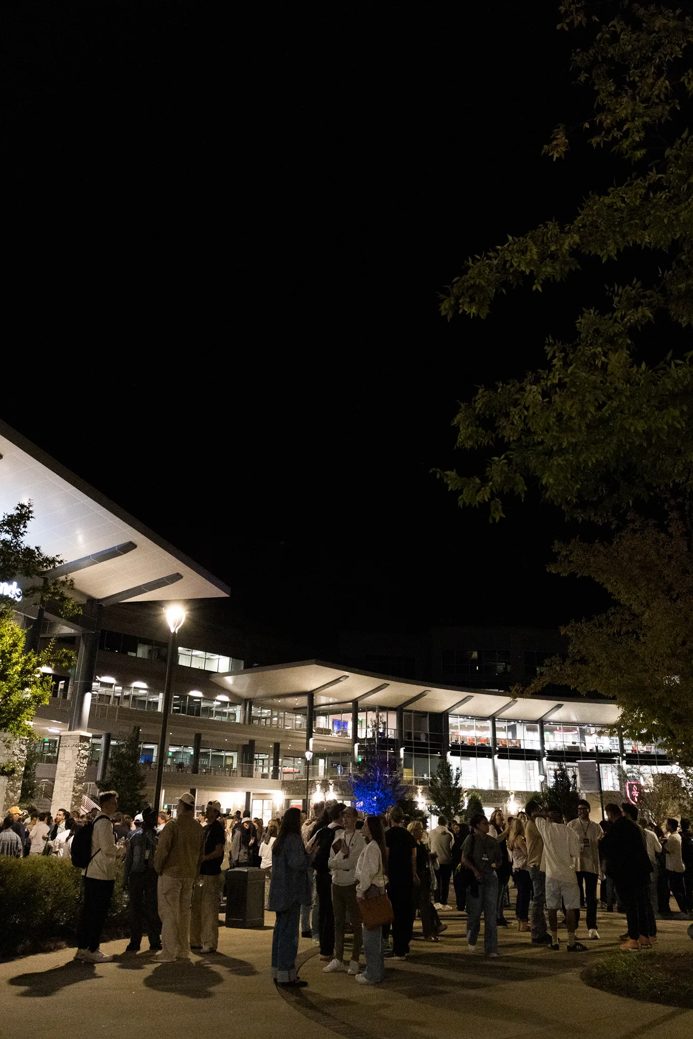 Crowd of people gathered outside a modern building at night, illuminated by streetlights and building lights, with trees on the right side.